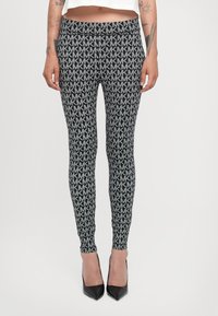 Leggings s uzorkom u crno-sivoj boji s ponavljajućim "MK" logotipima. Uski kroj s visokim strukom. Nošene s crnim visokim potpeticama.