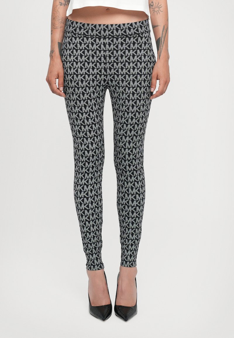 Leggings s uzorkom u crno-sivoj boji s ponavljajućim "MK" logotipima. Uski kroj s visokim strukom. Nošene s crnim visokim potpeticama.