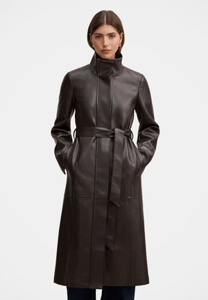 Femme portant un long manteau en cuir marron foncé avec un col montant et une ceinture nouée, debout, les mains dans les poches, sur un fond uni.