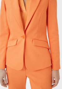 Helle orange maßgeschneiderte Blazerjacke mit passender Hose. Der Blazer verfügt über einen einzelnen Knopf, Reverskragen und strukturierte Taschen. Glattes Material.
