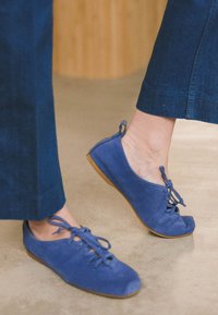 Blå mocka platta skor med rund tå, snörningar och en ljusbrun gummisula. Designen har en mjuk textur och minimalistisk stil.