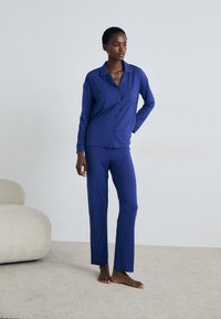 Violet pyžamo su ilgomis rankovėmis marškinėliais su kišenėmis, derinami su tiesiomis kelnėmis; pagaminta iš švelnios, lygaus audinio.