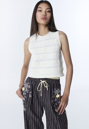 Giovane donna con capelli lunghi e lisci indossa una canotta bianca testurizzata e pantaloni neri a righe sottili con ricami floreali e vita con coulisse.