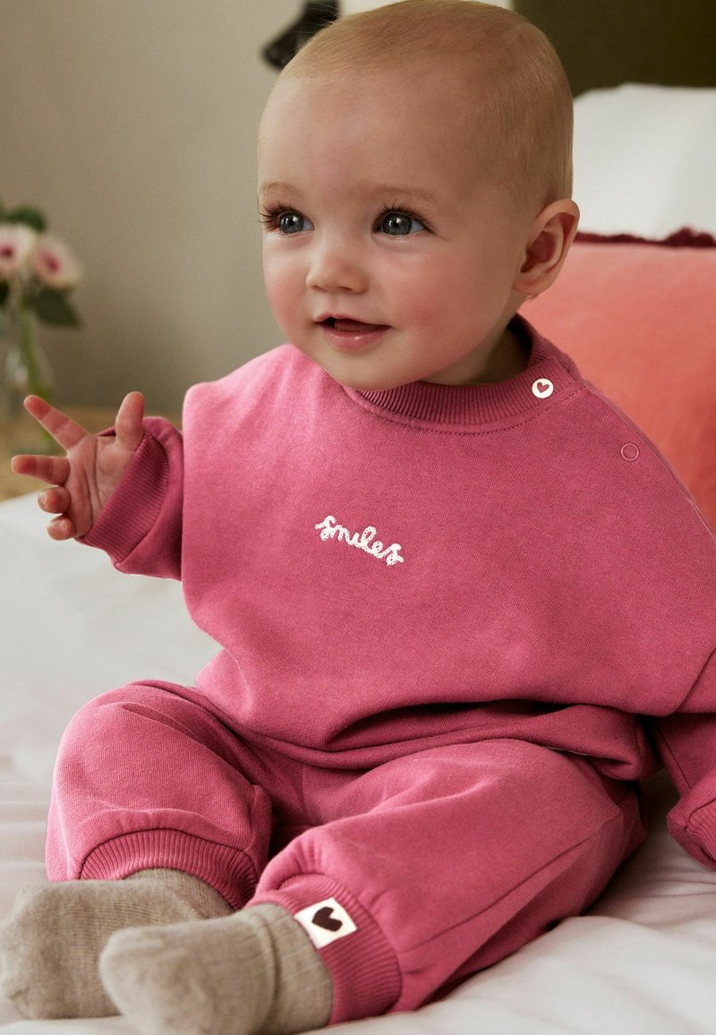 Rosa Sweatshirt und passende Hose aus weichem Stoff, mit gestickten "Smiles" und herzförmigen Akzenten am Bündchen. Einfaches Design.