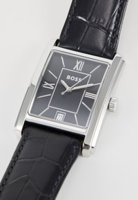 Montre en argent rectangulaire avec un cadran texturé noir, aiguilles argentées et chiffres romains. Bracelet en cuir noir avec un motif crocodile. Indicateur de date visible.