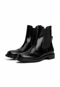 Bottines chelsea en cuir noir avec des panneaux élastiques sur les côtés et des détails brogue sur le bout. Texture lisse avec une semelle plate.