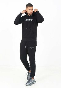 Conjunto de sudadera con capucha negra y pantalones deportivos, con un bolsillo frontal y cordones. La tela es suave y presenta un logo blanco en ambas piezas.