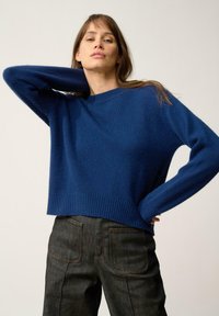 Niebieski, dziany sweter z prążkowanym dołem i mankietami, połączony z ciemnymi, teksturowanymi dżinsami z widocznymi szwami i tylnymi kieszeniami.