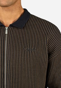 Pleteni zip-up pulover z rebrasto teksturo v rjavi in temno modri barvi. Ima raven ovratnik in diskretno vezenino logotipa na prsih.