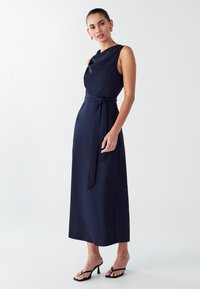 Navyblaues ärmelloses Kleid mit einem drapierten Ausschnitt, tailliertem Bund und einer langen, fließenden Silhouette. Trägt schwarze, riemenartige Sandalen.