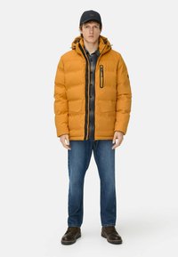 Gul polstret jakke med hætte, frontlynlås, sorte detaljer og en brystlomme. Matchet med en ternet skjorte og blå jeans.