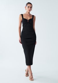 Vestido negro sin mangas con escote corazón, textura fruncida y largo hasta la rodilla, combinado con sandalias doradas de tiras.