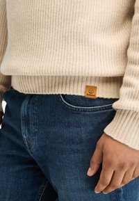 Maglione beige a coste con finitura strutturata, con toppe del logo marrone. Abbinato a jeans denim blu scuro.