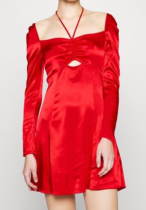 Kobieta w czerwonej satynowej mini sukience z długimi bufkami, marszczonym biustem, wycięciem pod biustem i ramiączkiem na szyję.