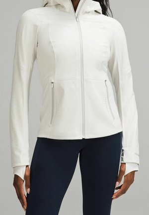 Strakke witte jas met capuchon en rits met duimgaten, gecombineerd met donkere leggings bij een staand persoon tegen een neutrale achtergrond.