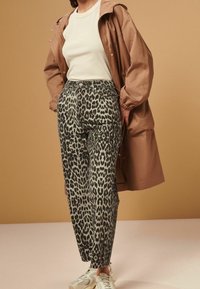 Person wearing beige tank top, brown oversized jacket, leopard print pants, and white sneakers standing against beige background.