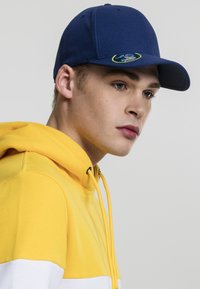 Cappellino da baseball blu navy con visiera curvata e una patch colorata. Indossato con un felpa gialla con strisce bianche e gialle.