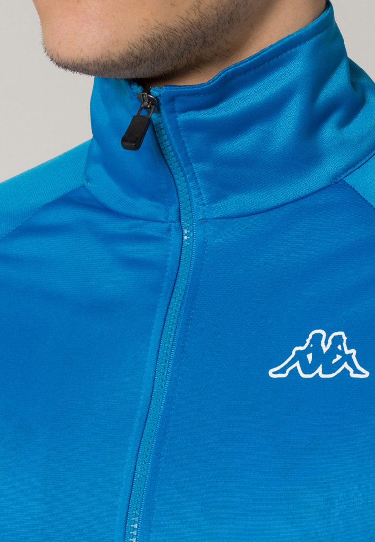 Giacca blu con zip e colletto alto, realizzata in tessuto liscio e leggero. Presenta un logo bianco sul petto e un tirazip nero.