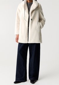 Faux fur coat in crème met een geknipte kraag, sluiting met één knop, zijzakken en een ontspannen pasvorm, gecombineerd met donkere denim wijde broek.