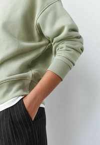 Gaiši zaļš džemperis ar ribotu aproci un apakšu, ar maigu tekstūru. Saskaņots ar melnām svītrainām biksēm, roka kabatā.