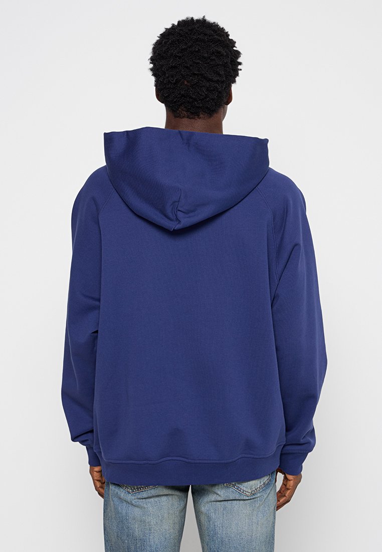 Granatowy hoodie z dużym regulowanym kapturem, wykonany z miękkiego materiału. Posiada luźny krój oraz ściągacze na rękawach i u dołu. Widoczne detale szwów.