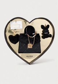 Mustast tikandiga käekott kunstkarva detailide, kuldsete riistvarade ja dekoratiivsete ripatsitega, mis asub südamekujulisest kastis läikiva kuldse sisemusega.