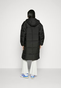 Melnā kapuce pūkainā jaka ar izšūtu dizainu, garā garumā un sānu kabatām, kombinēta ar baltām ribotām biksēm un daudzkrāsainām kedām.