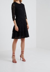 Robe en dentelle noire avec des manches trois-quarts, un ourlet superposé et un motif floral. Associée à des escarpins rouges à lanières.