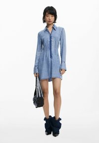 Vestido camisa de mezclilla con mangas largas, tejido azul claro, detalles de costura visibles, combinado con un bolso negro texturizado y botas oscuras.