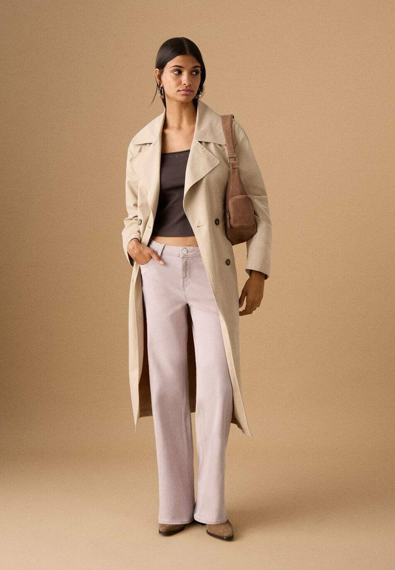 Trench beige sopra un top corto marrone, abbinato a pantaloni ampi rosa chiaro e tacchi beige. Piccola borsa marrone completa l'outfit.