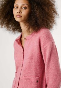 Een jonge vrouw met krullend haar draagt een roze cardigan met knopen en zakken, terwijl ze zelfverzekerd naar de kijker kijkt tegen een lichte achtergrond.