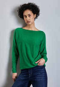 Grünes Langarmshirt mit einem Bootsausschnitt, das eine weiche Textur und enganliegende Ärmel aufweist, kombiniert mit dunkelblauen Jeans.