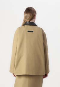 Khaki bunda s čiernym koženým golierom. Má uvoľnený strih a dva bočné švy. Na chrbte sa nachádza čierna nášivka na krku.