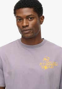 Camiseta de algodón de color morado claro con mangas cortas y cuello redondo. Presenta un logotipo gráfico amarillo en el área del pecho izquierdo.