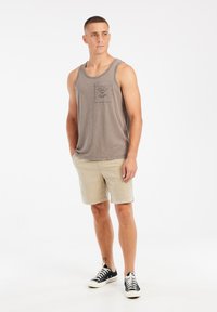 Grijze tanktop met een borstzak en bloemdesign, gecombineerd met beige shorts en zwarte sneakers, staande tegen een effen achtergrond.