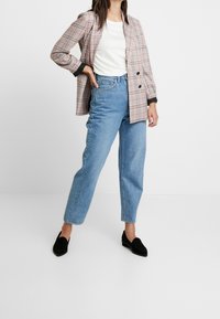 Karierter Blazer in Rot, Schwarz und Weiß, kombiniert mit einem weißen T-Shirt und hochtaillierten blauen Jeans. Schwarze Wildleder-Loafer runden das Outfit ab.