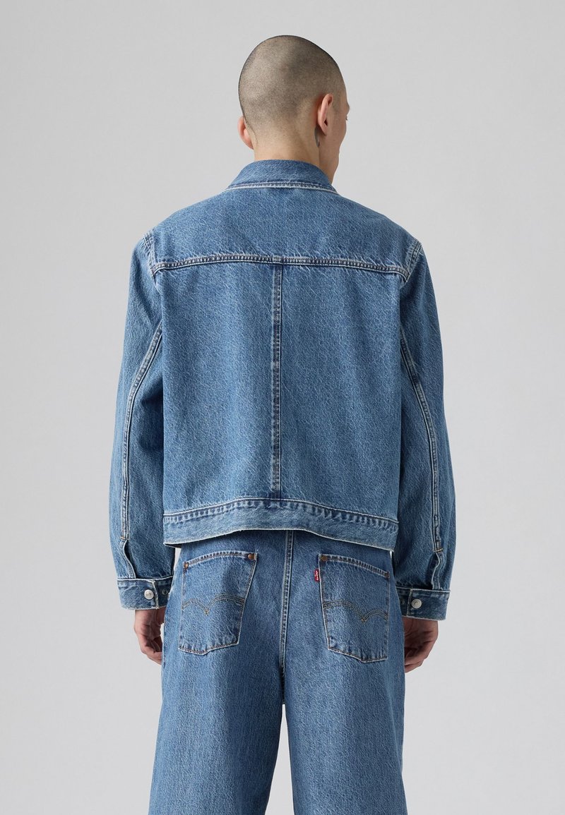 Giubbotto di jeans azzurro chiaro con colletto classico, maniche lunghe e due tasche posteriori. Presenta cuciture a vista e una vestibilità leggermente over.