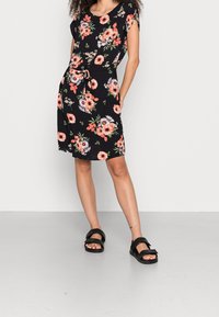 Blumenkleid mit schwarzem Hintergrund und pinken sowie weißen Blumen, kurzen Ärmeln und Taillengürtel. Tragen mit schwarzen Sandalen.