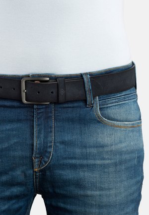 Crni remen s teksturiranim završetkom, s pravokutnom metalnom kopčom, nošen s plavim trapericama koje imaju ispucale i šivane detalje.