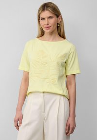 Żółta koszulka z krótkim rękawem z liściastym wzorem w ciemniejszym odcieniu, z okrągłym dekoltem i luźnym krojem, noszona z jasnymi spodniami.