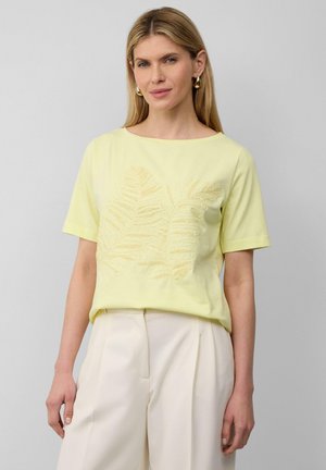 T-shirt jaune à manches courtes avec un motif de feuille dans une teinte plus sombre, doté d'un col rond et d'une coupe décontractée, associé à un pantalon de couleur claire.