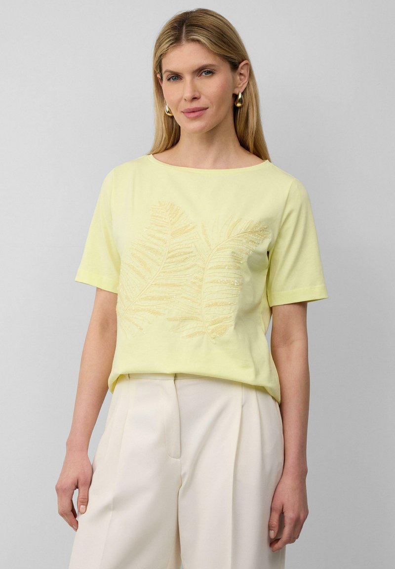 Żółta koszulka z krótkim rękawem z liściastym wzorem w ciemniejszym odcieniu, z okrągłym dekoltem i luźnym krojem, noszona z jasnymi spodniami.