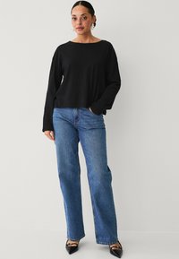 Top negro de manga larga con un escote amplio, combinado con jeans azul de tiro alto. La modelo lleva tacones negros de punta y pendientes blancos.