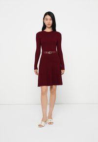 Donna in piedi che indossa un vestito lungo a maniche lunghe in maglia cable color bordeaux con una cintura abbinata e sandali con tacco aperto bianchi su uno sfondo bianco.