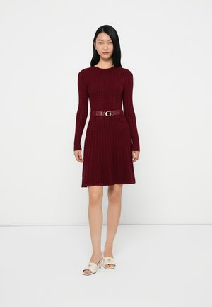 Donna in piedi che indossa un vestito lungo a maniche lunghe in maglia cable color bordeaux con una cintura abbinata e sandali con tacco aperto bianchi su uno sfondo bianco.
