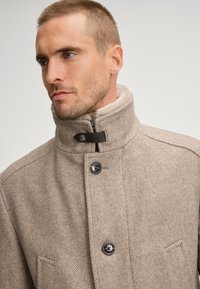 Jasnoszary o wzorze jodełki, z wysokim kołnierzem, czarnymi guzikami i miękką podszewką wewnątrz. Widoczny zamek błyskawiczny.