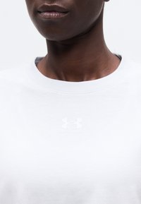 Εγγύς λήψη ενός ατόμου που φοράει λευκό πουκάμισο Under Armour με στρογγυλή λαιμόκοψη, δείχνοντας το λογότυπο και το κείμενο κεντραρισμένα κάτω από το λαιμό.