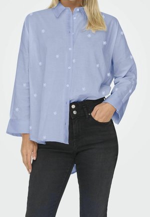 Camisa azul claro de algodón con botones, con bordado floral blanco, mangas largas y dobladillo curvado. Combinada con vaqueros negros.