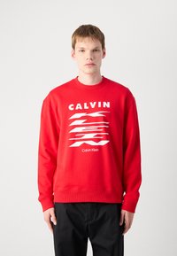 Sarkana kreklu izstrādājumi ar izteiksmīgu baltu "CALVIN" tekstu un abstraktu grafisko dizainu. Audumam ir gluda tekstūra ar ribotiem piedurknēm un apakšu.