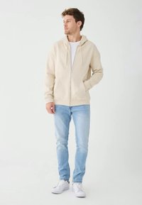 Giovane in piedi, indossa una felpa beige con zip, t-shirt bianca, jeans azzurri e sneakers bianche, guarda verso sinistra.